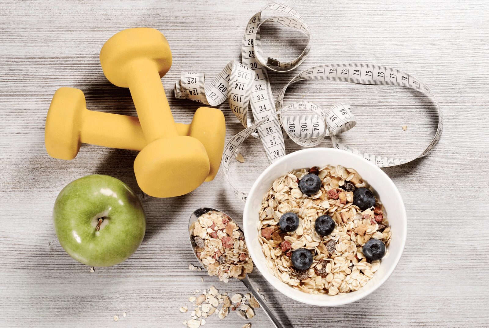 Oats, fresh fruit, light hand weights and a tape measure arranged on a wooden surface